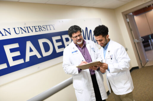 A male doctor of chiropractic and chiropractic student in white coats look at a clipboard and discuss how to become a chiropractor