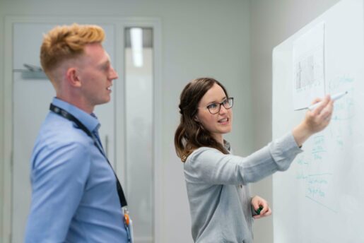 A male looks on as a female writes on a white board